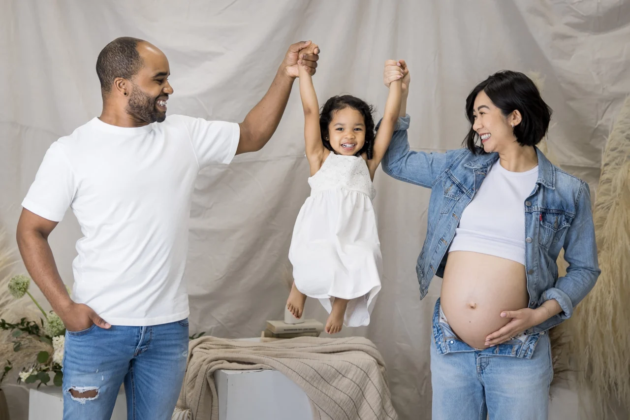 Little girl being swung by the hand by parents as part of studio maternity photo session by Edmonton portrait photographer Paper Bunny Studios