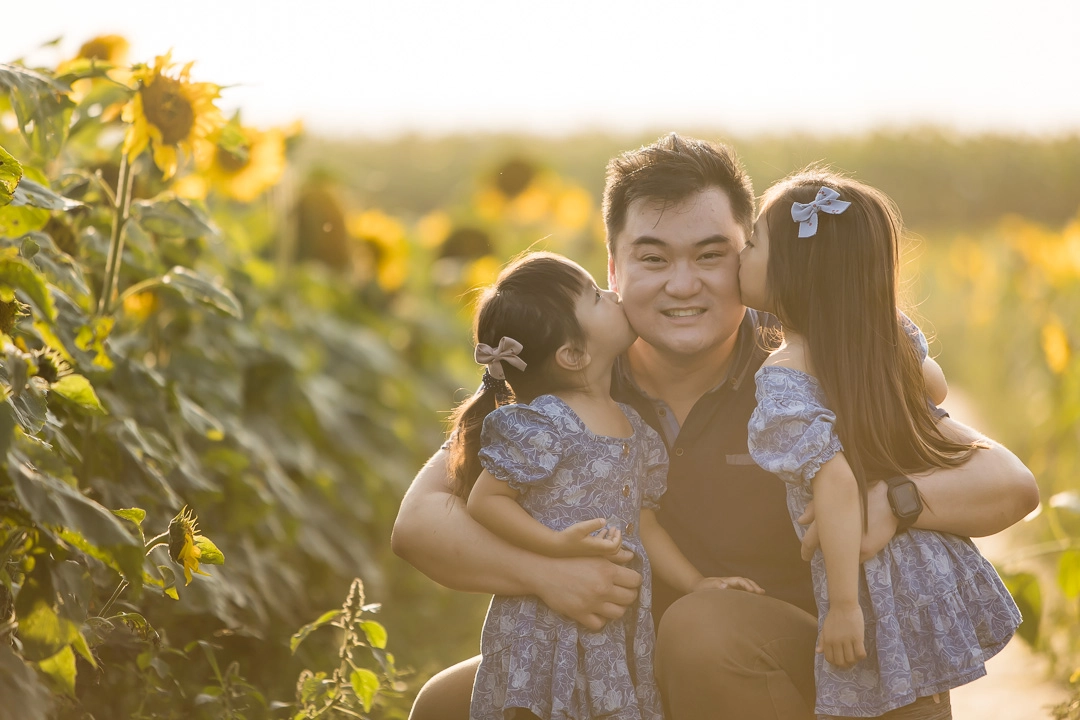 Edmonton family photos in sunflower field of daughters kissing dad on cheek by fine art family photographer Paper Bunny Studios