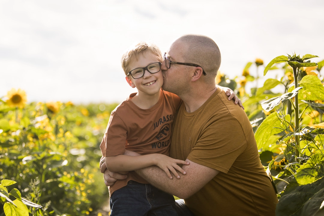 Edmonton family photos in sunflower field of dad kissing son on cheek by fine art family photographer Paper Bunny Studios